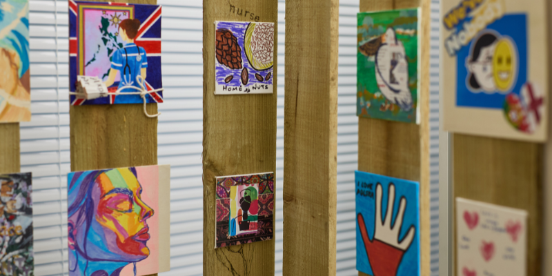 Close-up of wooden panels display colourful, square art pieces, including a vibrant face, a hand a nurse looking at a painting of a map, nuts, a bird and more.