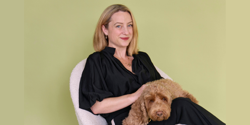 Christie Watson, a white woman with blonde hair and blue eyes, is wearing a black blouse and trousers. She sits on a white chair against a light green-yellow background, smiling gently, with a fluffy brown dog resting on her lap.