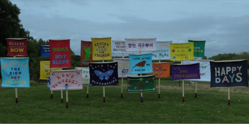 Photograph of several placards, made from colourful fabric, in a field. They feature messages such as "adjust your sails for change" and "we are treasures".