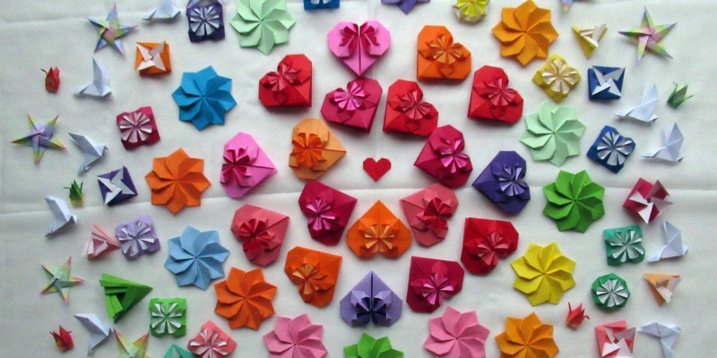 A vibrant array of origami flowers, hearts, stars and birds in various colours including pink, blue, red, purple, yellow, green, and orange, arranged in a circular pattern on a white sheet.