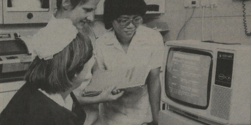 Greyscale image from 1985 of three women nurses in a hospital room, all looking a computer screen, discussing and ordering hospital supplies digitally.