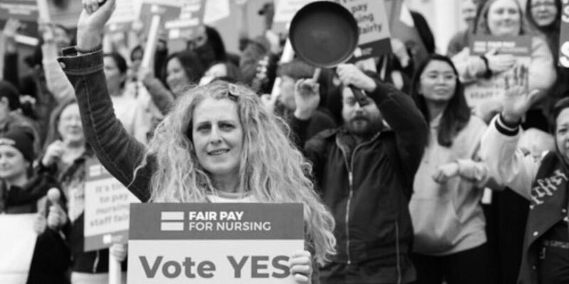 Member of nursing staff on picket lines holding Fair Pay for Nursing "Vote Yes" sign.