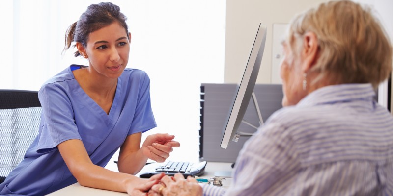 Woman nurse in blue scrubs sitting at a desk speaking to person and holding hand