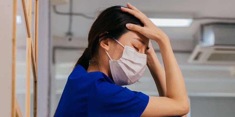 Nurse, female and east Asian, wearing face mask and royal blue uniform holds head in hands as if in distress