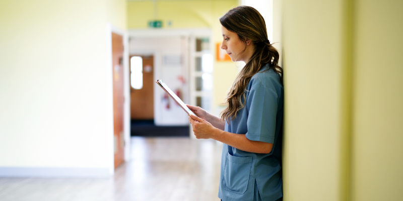 Female nurse stands with her back against a wall in a hospital hallway, reading notes on a clipboard