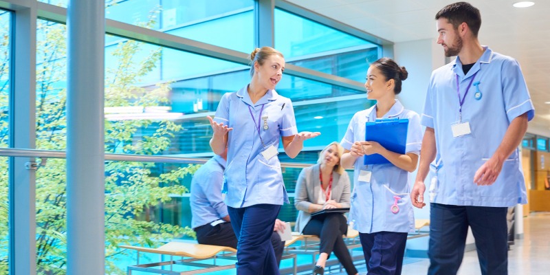 Three members of nursing staff are shown walking inside a hospital. There is a large glass window next to them flooding the room with light. They are all wearing navy trousers and light blue uniforms.