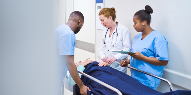 Two members of nursing staff and a doctor speak to a patient who is lying flat on a bed in a hospital corridor. The nursing staff wear blue uniforms. One is male and has his back to the camera, while the female nurse holds a clipboard. The doctor has a white coat and stethoscope