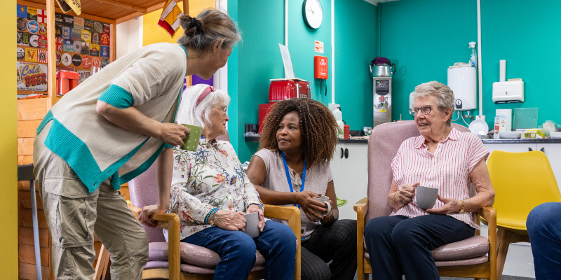 A retired community of friends are shown relaxing together in a common space alongside health care carers. One of the ladies has dementia, and is receiving support for it. Two of the women sit in seats, one stands next to them holding a mug and another crouches down to talk to them. Brightly coloured walls with notices pinned on them are seen in the background.