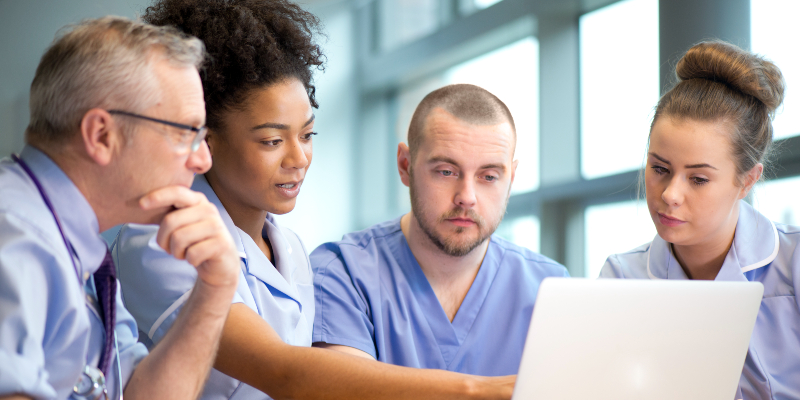 Four medical staff are seen in a meeting. They are sitting at a table and all looking at the same laptop screen. From left to right there is a middle-aged man wearing glasses, a young black woman pointing at the screen, a young white man with short hair and a beard, and a young white woman. The three on the right appear to be nursing staff in light purple uniforms.