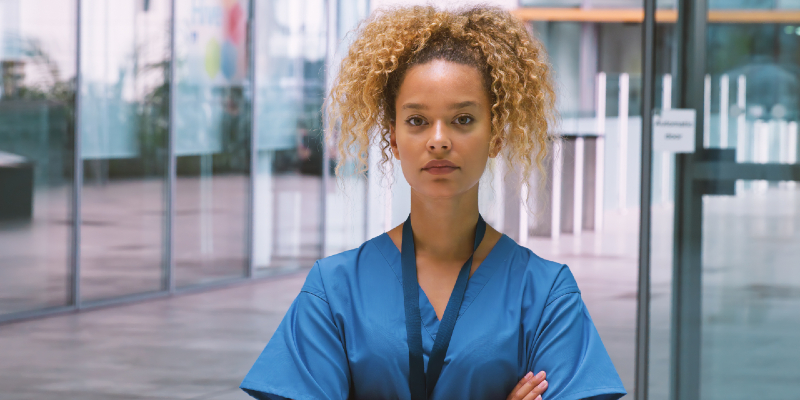 Female nurse wearing scrubs standing in a hospital with her arms crossed and looking serious 