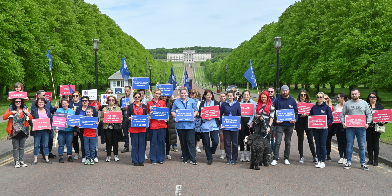 A group of nursing staff are shown holding placards marching with Stormont in the background