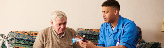 male community nurse chatting with an elderly patient in their home. the nurse is showing the patient how to use an inhaler with spacer
