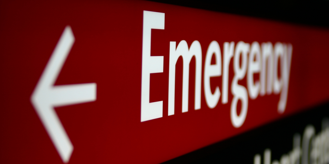 An emergency sign is seen in close up, with white writing on a red background and an arrow in the foreground of the picture.