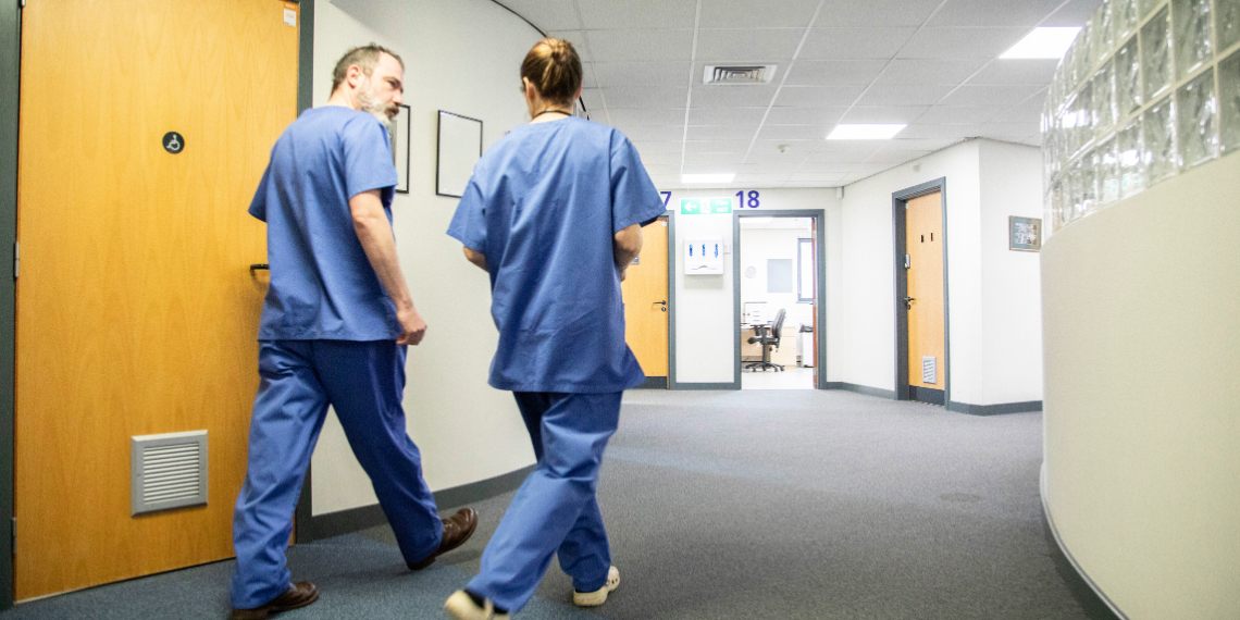 Two members of nursing staff walk down a corridor. One is male and one female. They both wear blue uniforms and are walking away from the camera, from the left to the right. Wooden doors can be seen off the corridor in the foreground and background, as the corridor bends around to the right