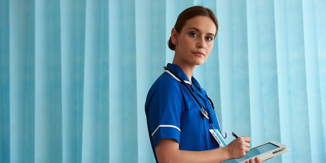 A health care worker stands looking into the camera. She is wearing a blue uniform with short sleeves and behind her is a light blue curtain. She holds a computer tablet and pen or stylus which she is holding to the screen. She has dark brown hair which is tied up.
