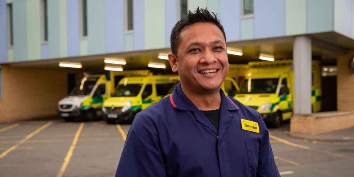 A nurse stands outside a hospital, seen from the waist up, with ambulances parked in a bay visible behind him. He is wearing a navy uniform with red trim and has a yellow name badge on. He is smiling, and looking off camera.