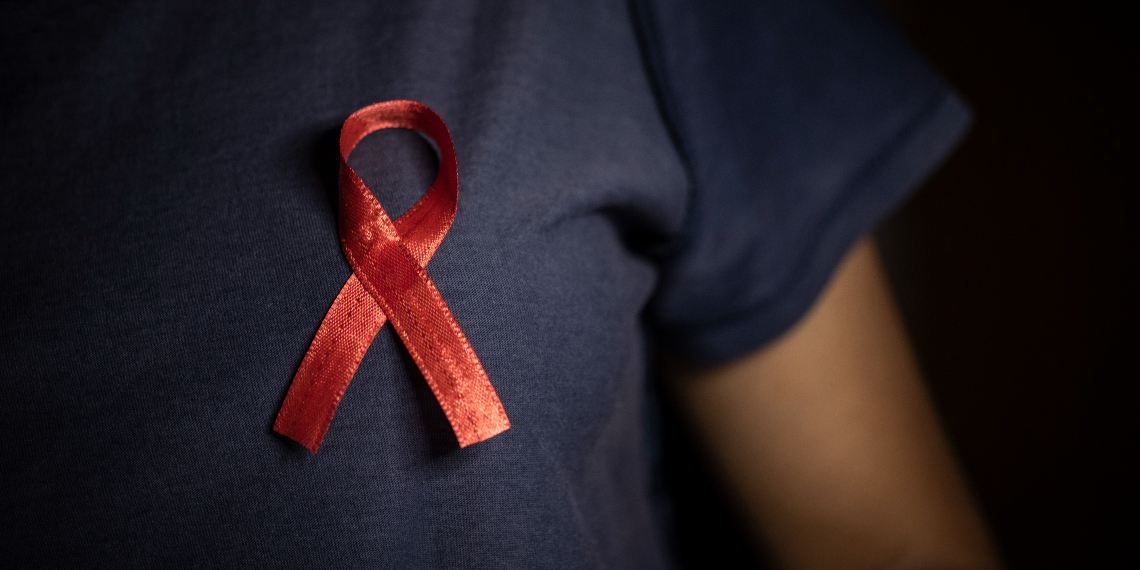 A close up shows a red World Aids Day ribbon pinned on a person's dark blue nursing uniform
