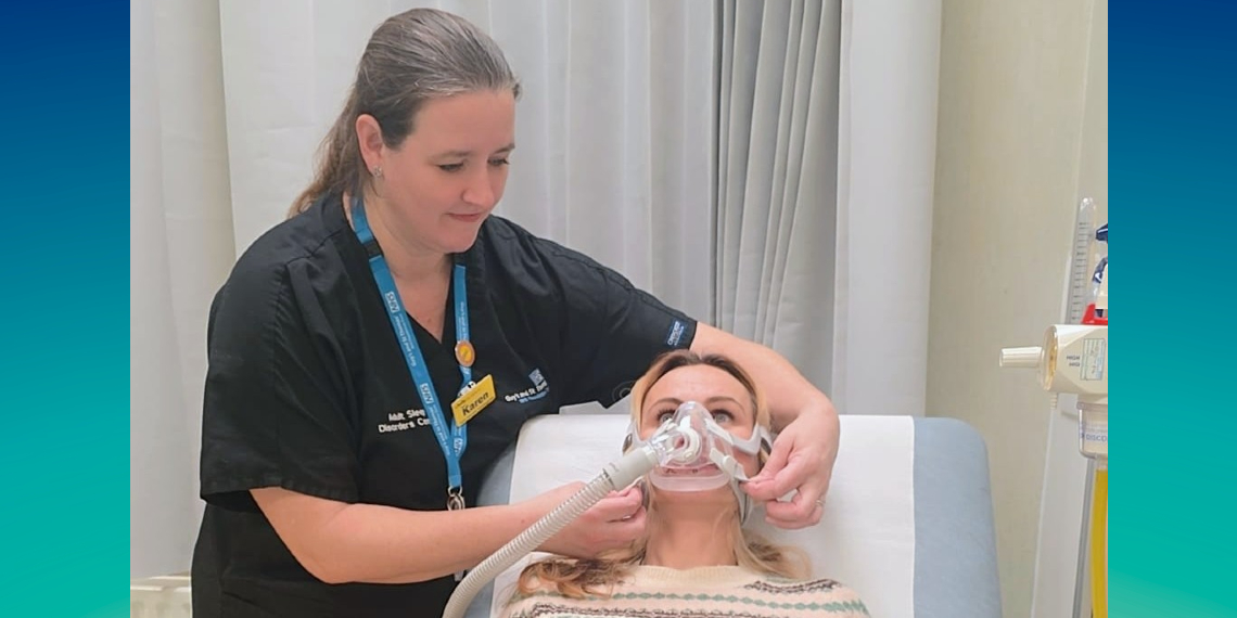 A nurse in a dark blue uniform fits a sleep CPAP machine onto a patient who is lying on a bed. The machine fits over the mouth and nose and has tubes coming out of it. A hospital curtain is visible in the background. The image has a green/blue border on the right and left side.