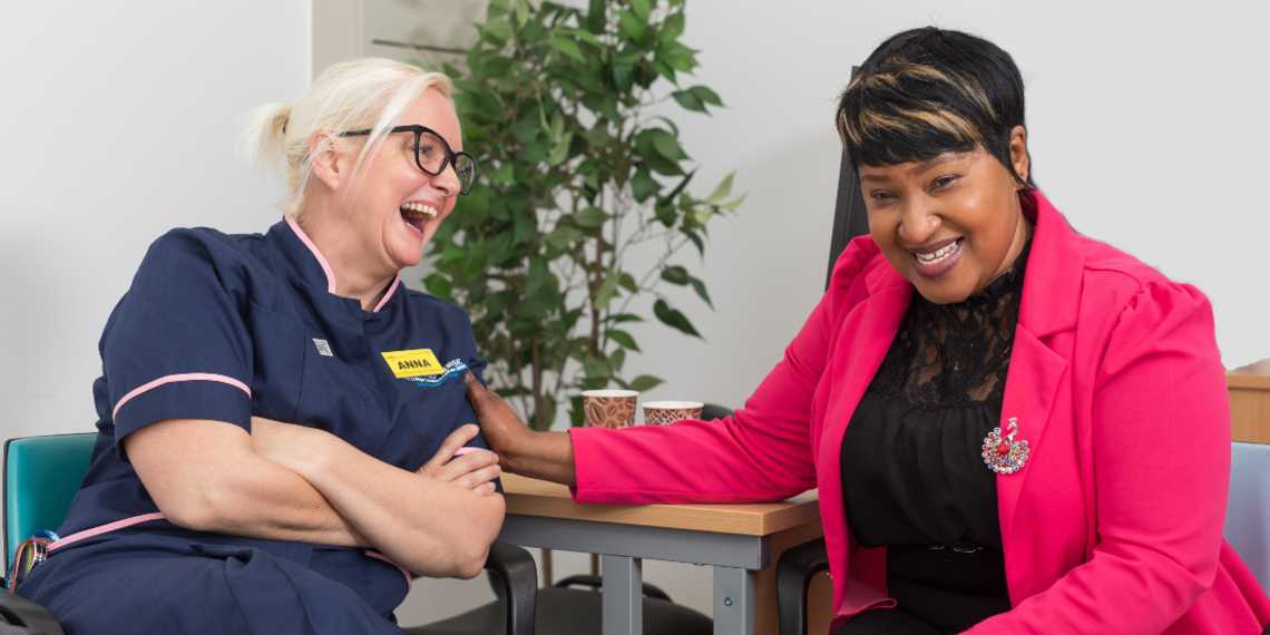 Two nurses sit at a desk facing each other. Anna, on the left, wears her navy nurse's uniform while Nima, on the right wears her own clothes, a bright pink jacket and black clothes. They are good friends, and are laughing together while drinking tea.