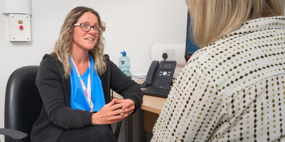 Kelly, a mental health nurse, sits at her desk facing a patient. Kelly is wearing a blue top with a black cardigan over the top. She has glasses and dark blonder hair. She is steepling her fingers and her body language is open as she listens to her patient.