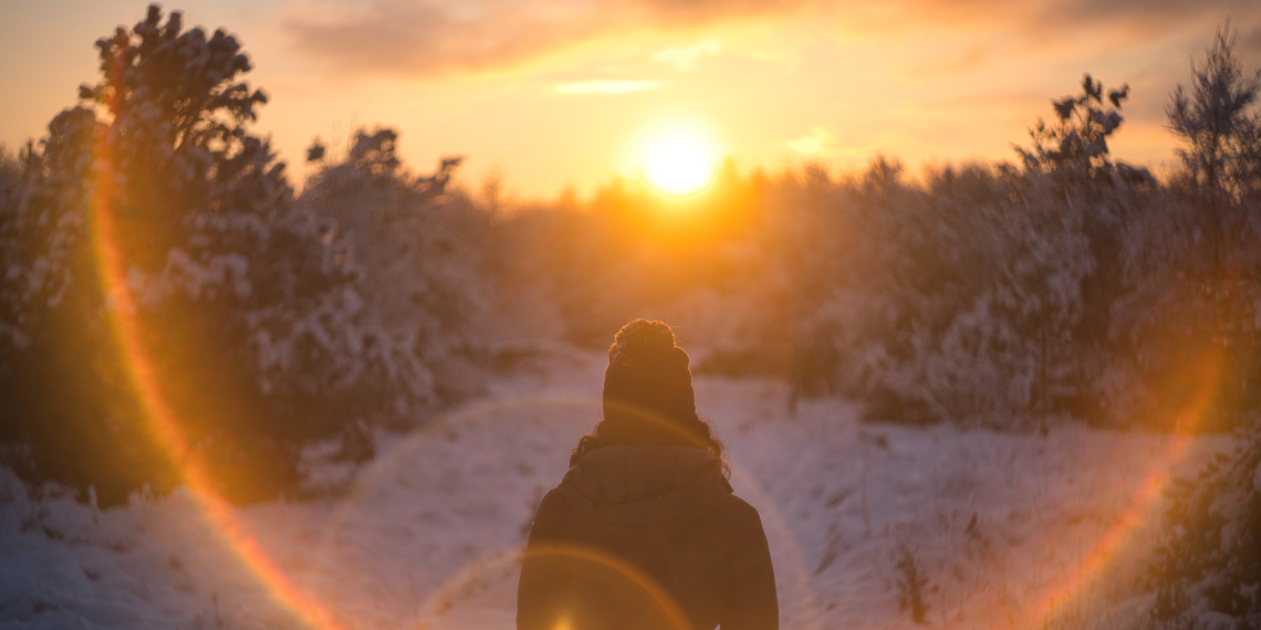 A woman wearing a winter coat, with her back to the camera, walks in snowy woodlands facing an orange rising sun
