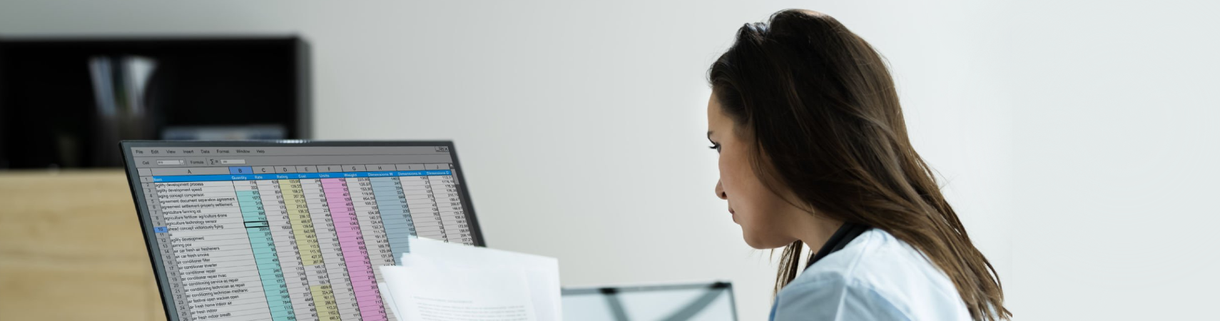 Nurse at desk at computer, looking at paperwork and spreadsheet