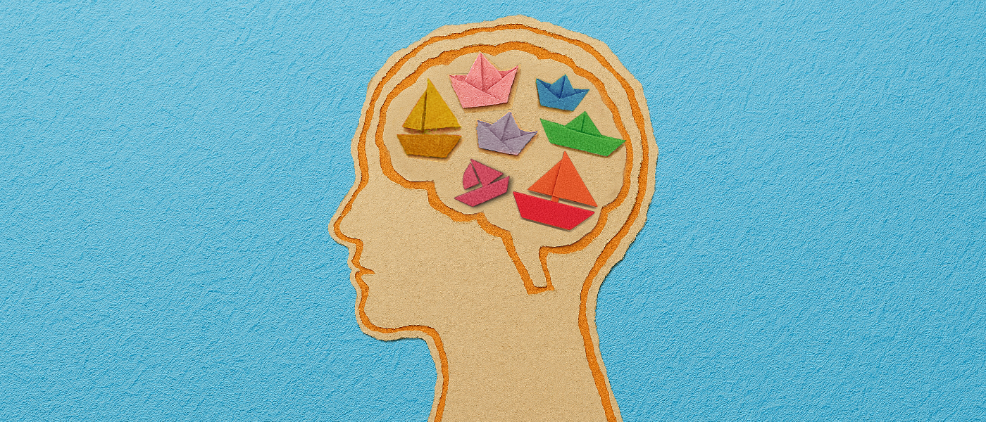 A scrapbook-style illustration of a human head in profile, with the brain area filled with small, folded paper boats in a variety of bright colours.