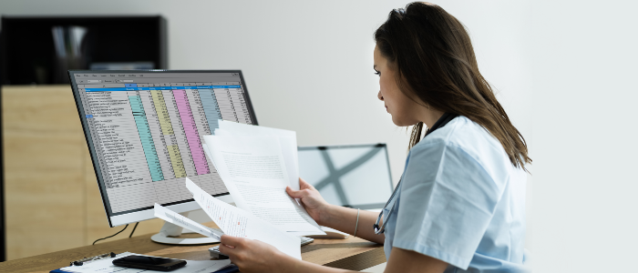 Nurse at computer, trying to make sense of paperwork and a spreadsheet
