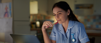 Female nurse sat at her kitchen table in her home, looking at her laptop deep in thought