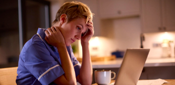A tired-looking nurse in scrubs sits at a kitchen table, staring at a laptop screen with a hand on her forehead. A mug, phone, and stethoscope lie nearby, suggesting she's working late or dealing with stress.