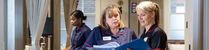 Three nurses in uniform inside a healthcare setting, with two discussing notes from a folder while another works in the background.