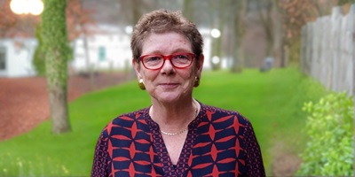 A bust portrait of Professor Anne Whittaker, a white lady with short, light brown hair, blue eyes who is smiling faintly at the camera. She is wearing red glasses, gold earrings and a necklace, and a blue and red patterned blouse. 