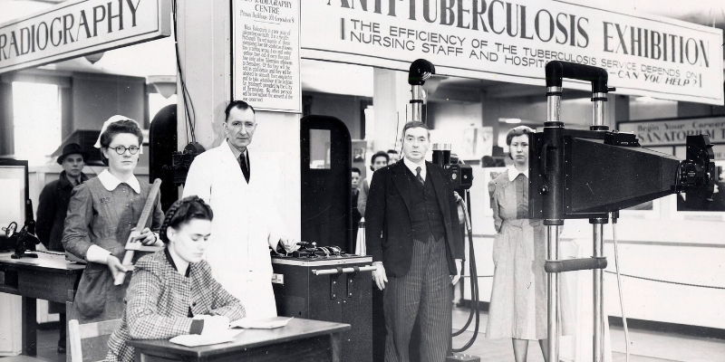 Greyscale image of medical staff, including nurses, at a Tuberculosis sanatoria and public health communication event. Four members of staff are standing looking at the camera, surrounded by information boards and various equipment. A fifth individual sits at a desk writing near to the camera.