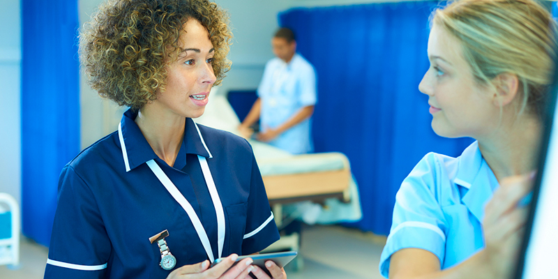 A nurse talks to a junior nurse with a third nurse in the background preparing a bed
