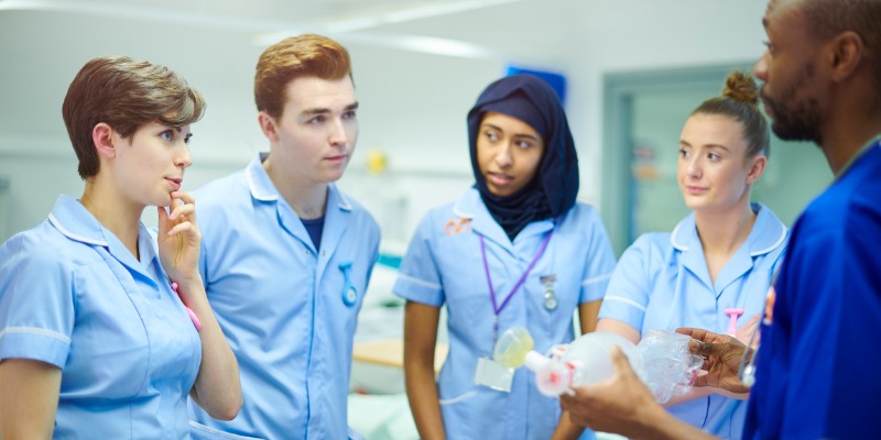 A group of nurses stand in a hospital ward and listen as another nurse speaks to them
