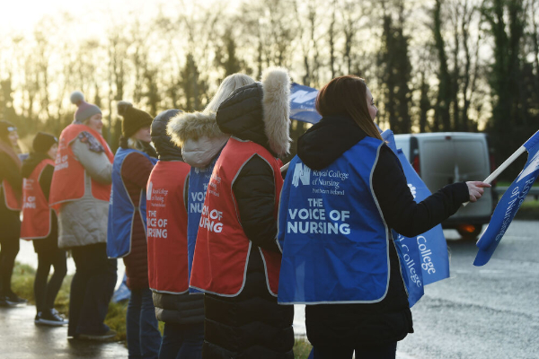 Nursing staff and supporters wearing campaign tabards stand beside a roadside, holding flags during an outdoor demonstration.