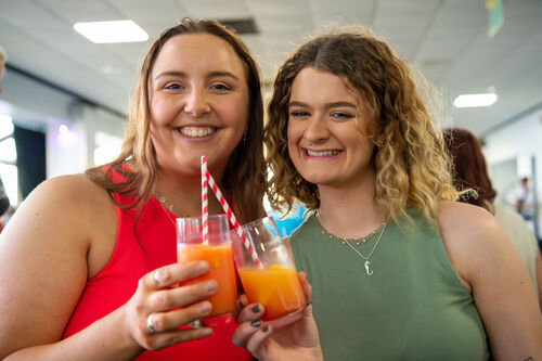 Two smiling women holding glasses of orange juice with straws, posing together at an event.