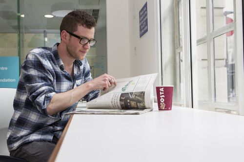 A man sits at a table next to a window. He is drinking a coffee and reading a magazine 