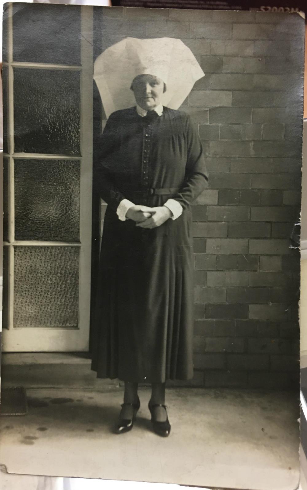 Photograph of an older nurse in dark uniform (possibly a sister) taken outside hospital buildings c1920. Larger hats like this were often worn by matrons as a sign of status.