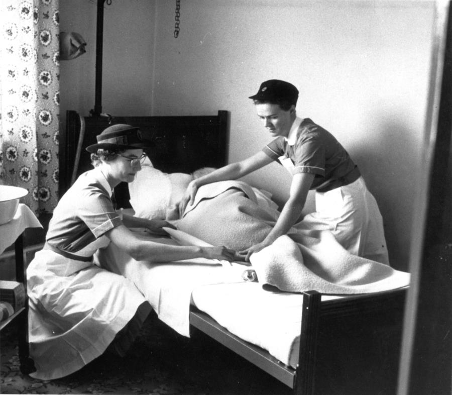 Two district nurses move and prepare a bed-bound older patient for bathing, c.1955-1965. RCN Archive. 