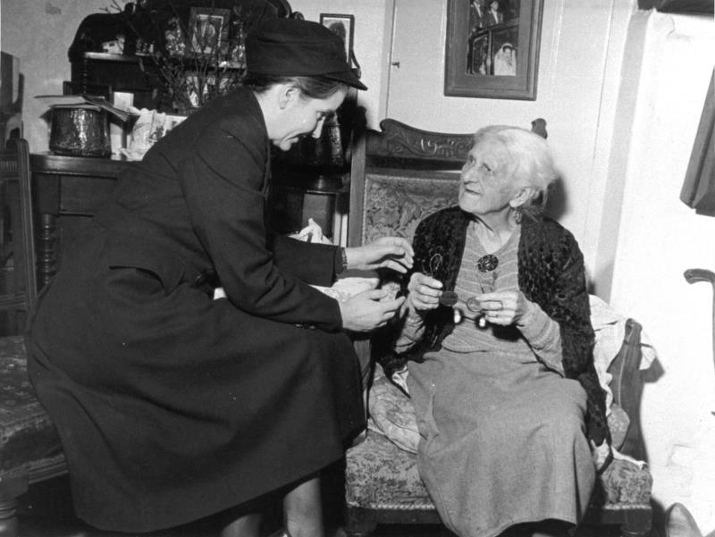 Black and white postcard with a district nurse attending an elderly person in their home. 