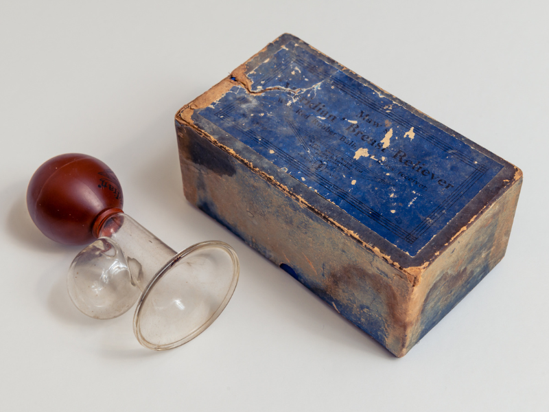 historical glass breast pump placed next to its original box. The box is blue and well worn.