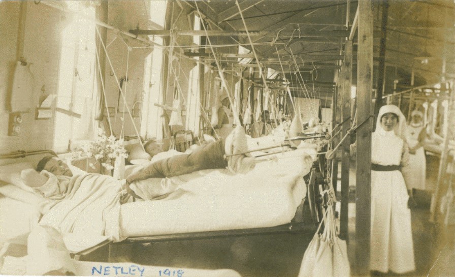 Black and white photograph of a ward with a row of patients in beds with various limbs suspended from pulleys on the left and a nurse in the foreground on the right