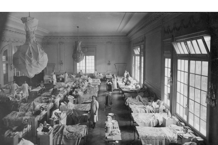 Black and white photograph of a room taken from aerial view filled with patients in beds, nurses and a chandelier wrapped in a sheet hanging from the ceiling. 
