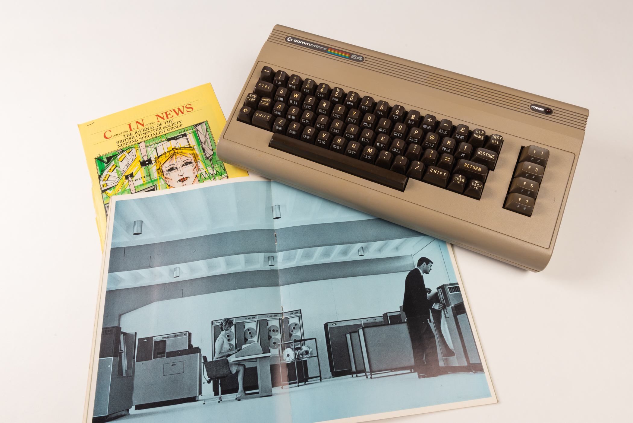 A beige, vintage keyboard with dark brown keys alongside a yellow magazine with an angular line drawing of a nurse on a green background, and an open booklet showing an old, blue-tinted image of two people in an office full of computer mainframes. 