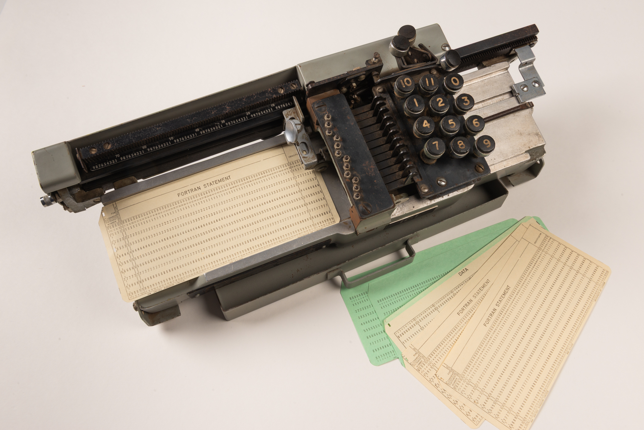 A large metal device with a typewriter-style keypad featuring numbers 0 to 11. The keypad is mounted on a rail allowing it to slide across the device as the user types. Placed on a tray to the left of the keypad is a card showing horizontal rows of numbers. 