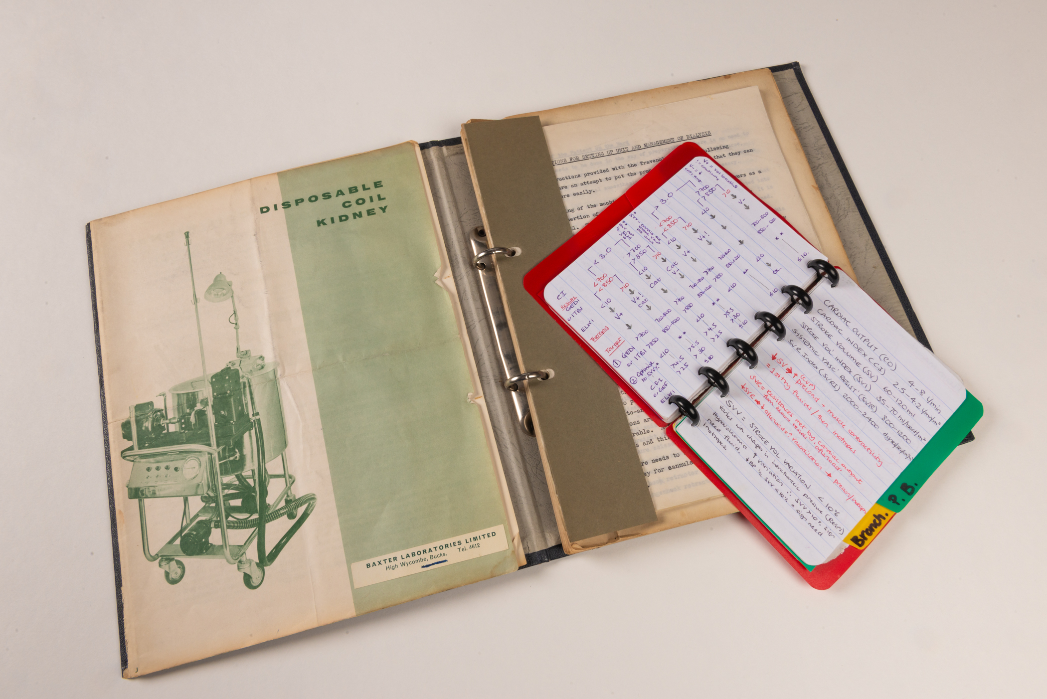 An open, ring-bound folder showing a sepia and green image of an old dialysis machine with the words Disposable Coil Kidney in green above. Covering the other page of the folder is a small, open notebook with a red cover showing technical notes. 