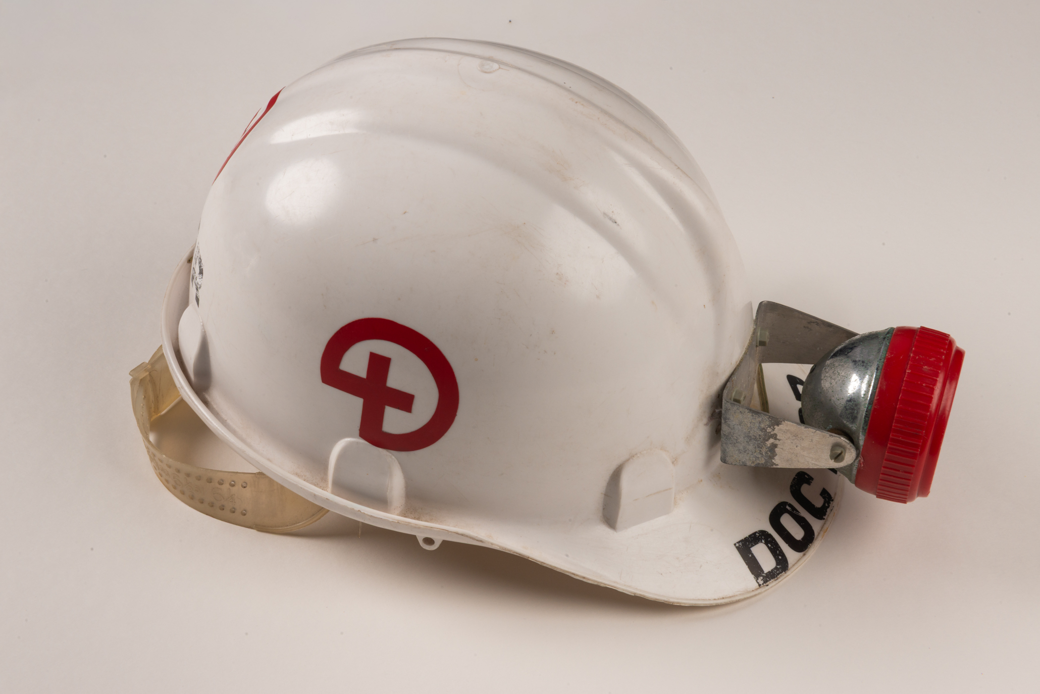 A white hardhat with a red cross in a circle on the side and a metal lamp with a red rim affixed to the front. The first letters of the word doctor are visible beneath the lamp on the helmet's visor. 