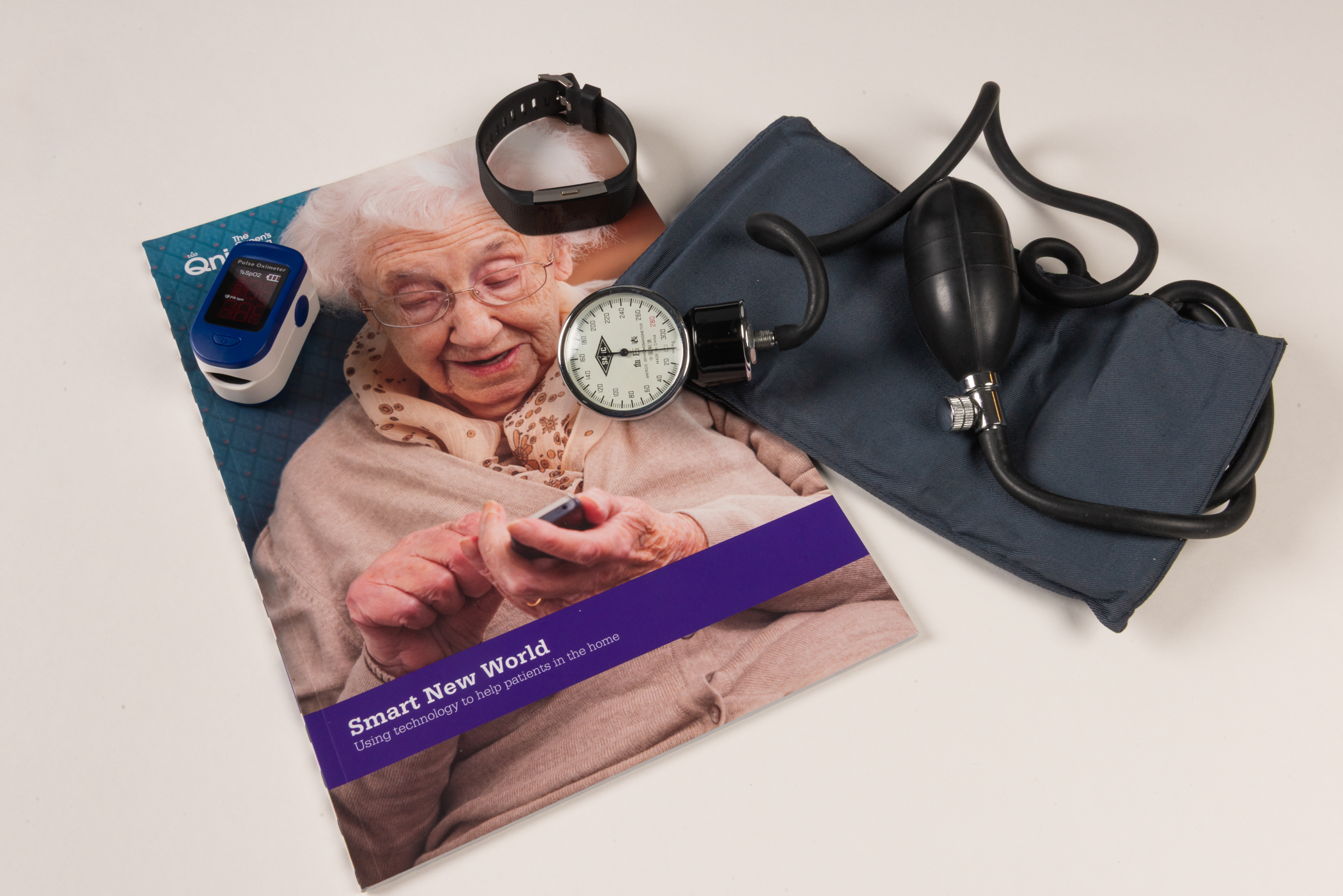 A small white and blue device with a digital display, a watch with a black strap and a blank, glass screen, and a navy blue arm band attached to rubber tubing with a hand pump on one end and an analog dial on the other. All of these are arranged atop a magazine cover showing an elderly woman sat using an old mobile phone.