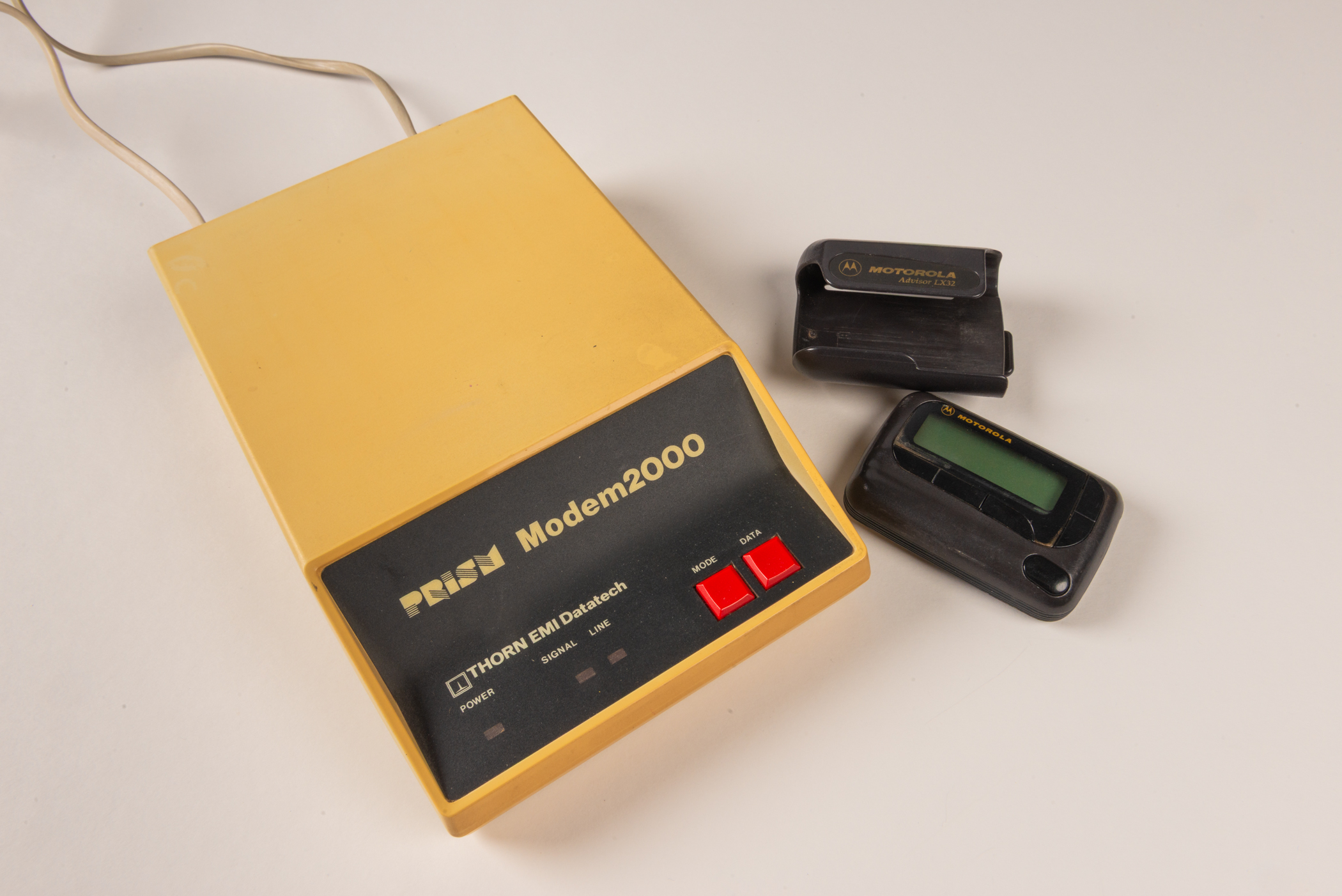 A small, black, rectangular pager with one button and a digital display beside a larger, yellow plastic box with a concave, black face at one end featuring two red buttons for mode and data and three lights for power, signal and line. 